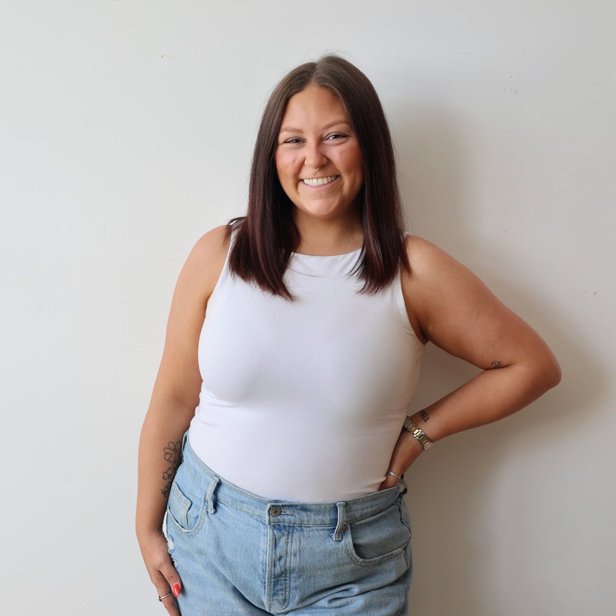 A woman with shoulder-length brown hair smiling, wearing a white sleeveless top and light blue jeans, standing against a plain off-white wall.